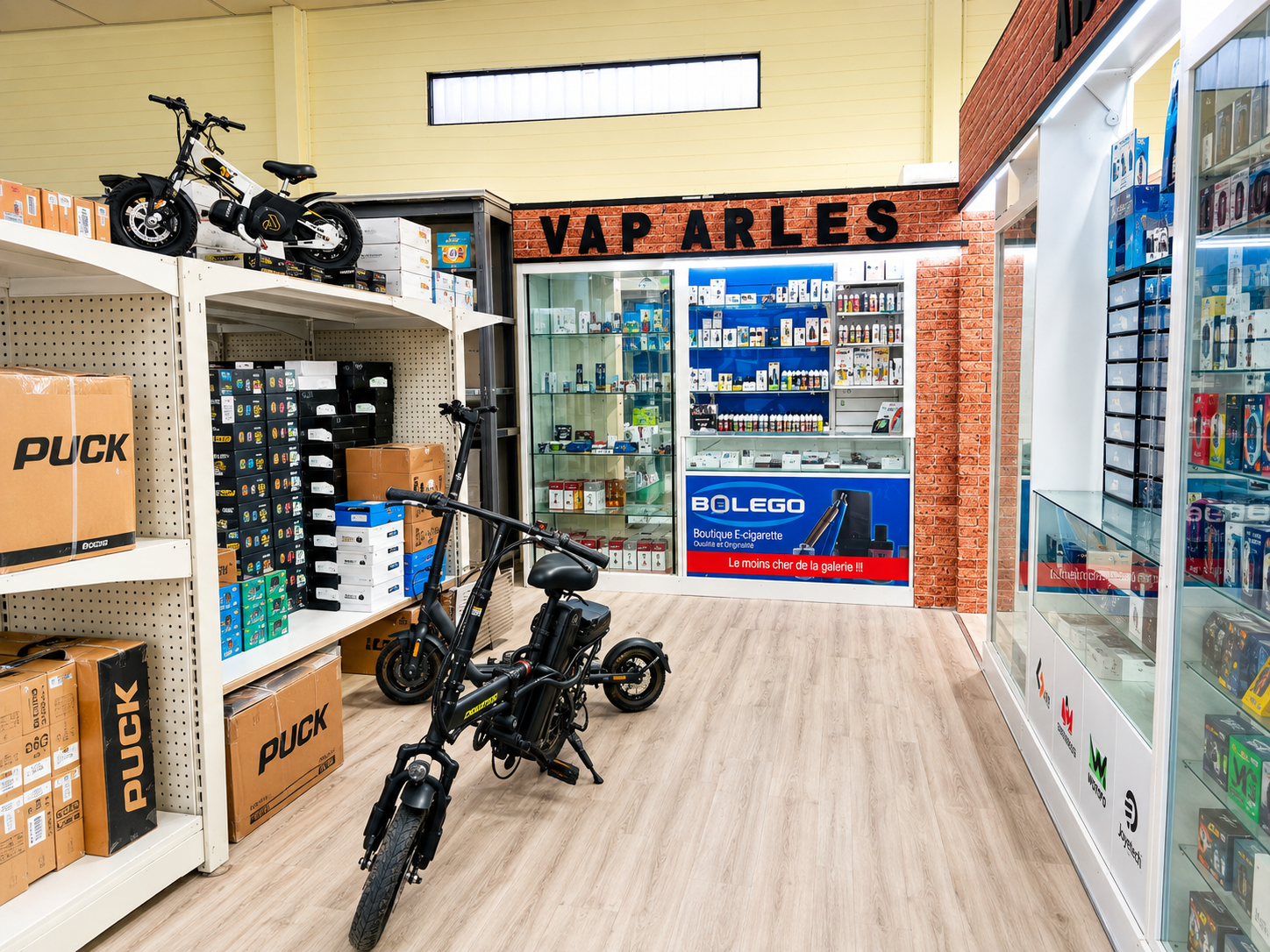 Allée commerciale avec plusieurs vélos électriques, des rayonnages de cartons et l’enseigne VAP ARLES